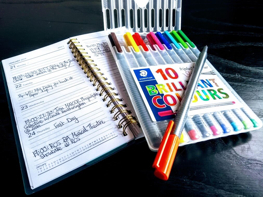 A diary lying open with appointments written in it in black pen, a case of 10 brightly coloured pens open with the orange out ready to be used.