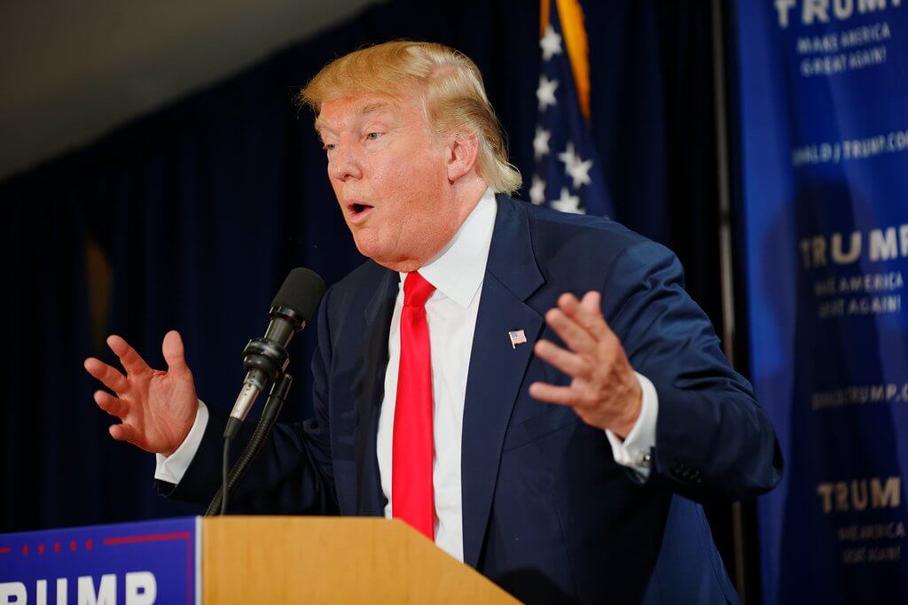 Donald Trump mid-speech at a rally, dressed in a suit and tie, hands gesturing expansively.