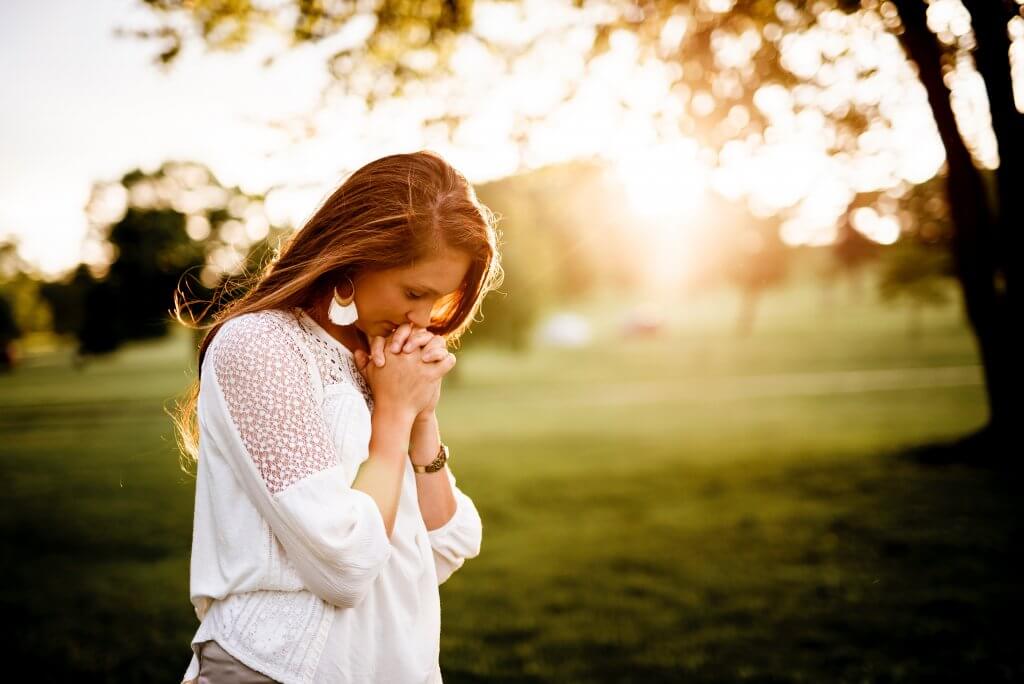A woman praying/looking down contemplatively at her clasped hands, standing outside in the sun.