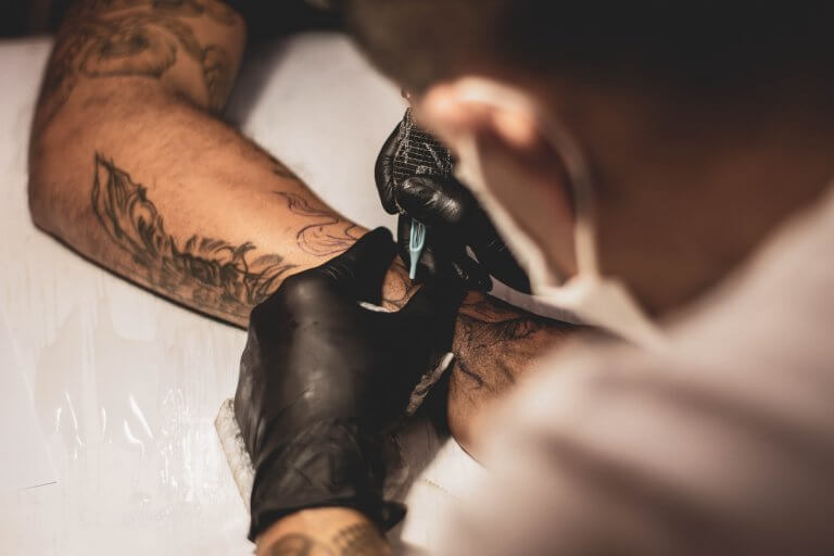 A tattoo artist with black gloves on is inking the arm of a person who already has a few tattoos.