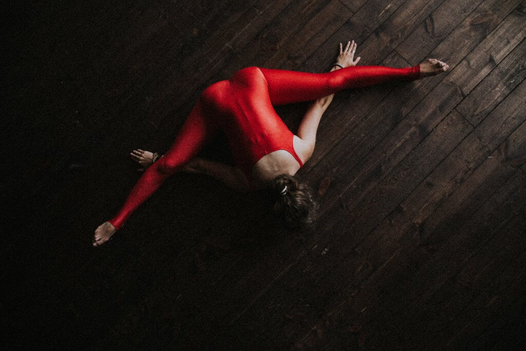 A woman sitting on the floor, stretching her legs out wide, her arms stretched out wide beneath them, palms down. She wears a bright red leotard/outfit.