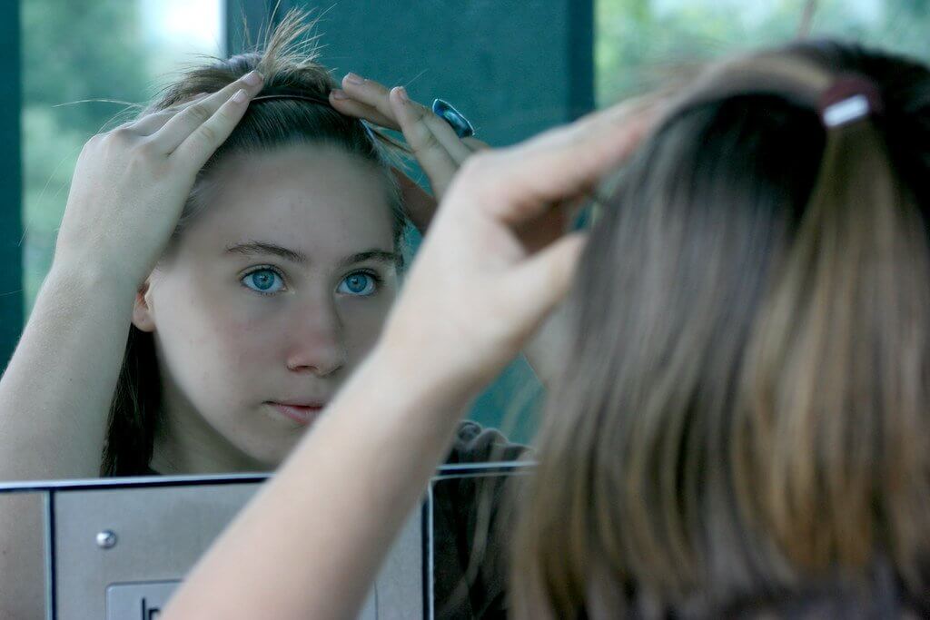 A teenage girl looks doubtfully at herself in the mirror as she pushes a hairband back through her hair.