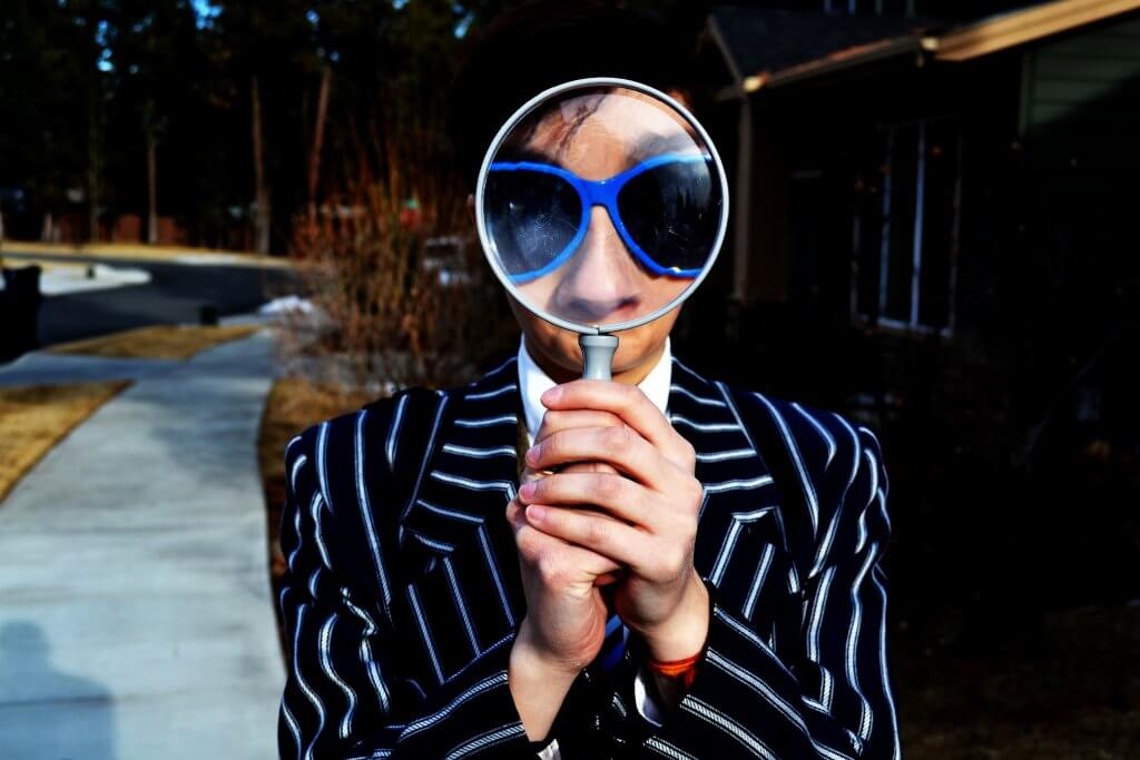 A man in a pinstripe suit and blue-rimmed sunglasses holding a magnifying glass up to his own face, making his eyes and nose seem disproportionately big.