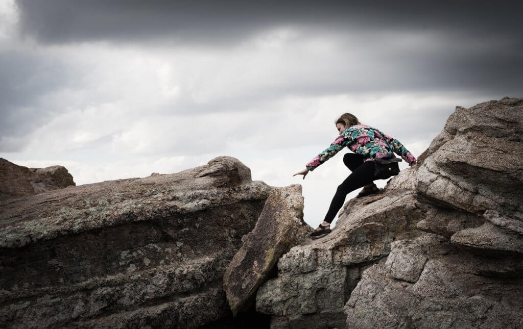 A woman tentatively pushing off her safe rock to risk reaching another rock.