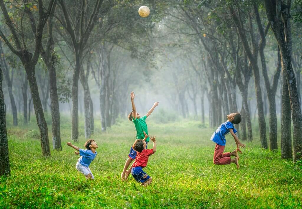 A group of young boys play with a ball in a clearing in some woods, jumping to catch the ball.