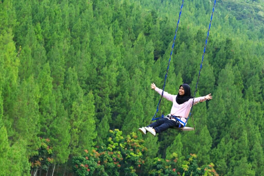 A young Muslim woman sitting on a swing high above a forest, arms open wide, smiling.